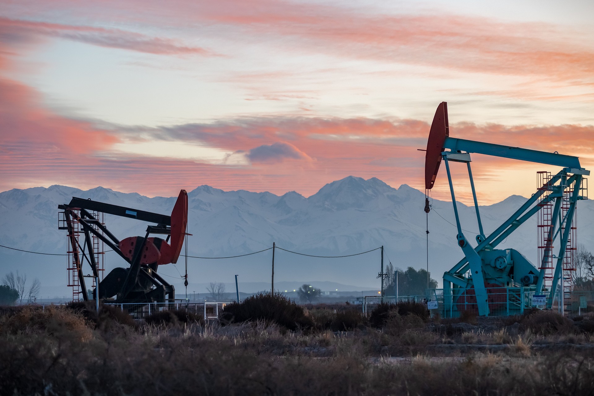 Oil pumping system from a well with silhouettes of electric poles and power lines against snowy mountains in a red sky.